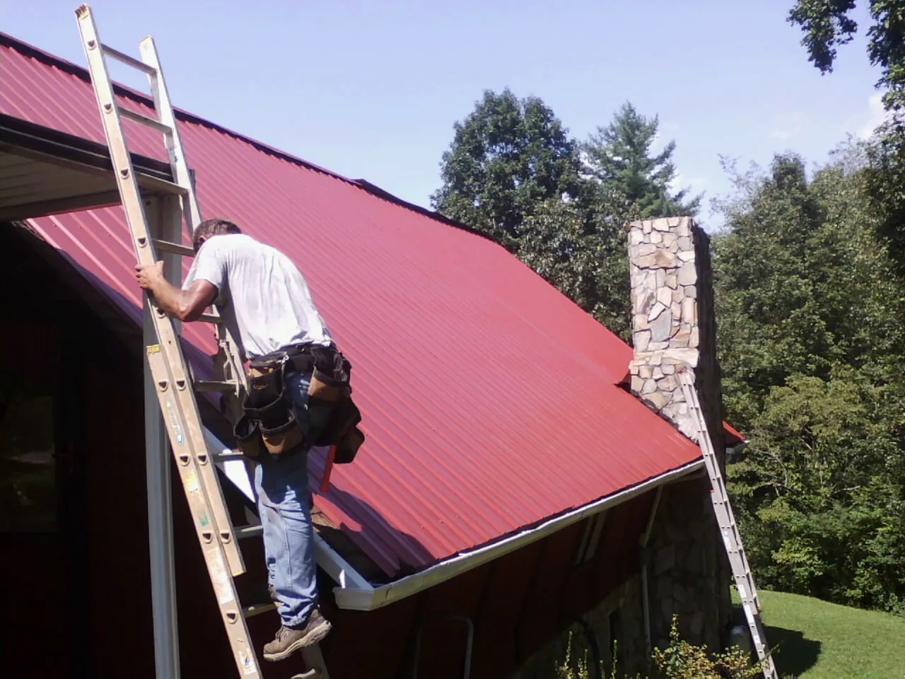 Roofer on a ladder beside a red metal roof and stone chimney — Soffit & Fascia Repair by Roofing Brothers in Jermyn
